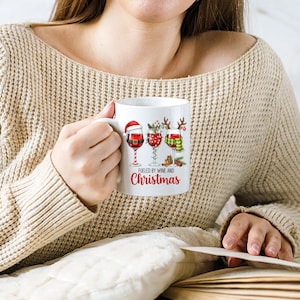 Peut inclure: Mug en c&eacute;ramique blanche avec un motif de No&euml;l. Il pr&eacute;sente trois verres &agrave; vin d&eacute;cor&eacute;s avec des &eacute;l&eacute;ments festifs, dont un bonnet de P&egrave;re No&euml;l et une &eacute;charpe. Le texte "Fueled by Wine and Christmas" est imprim&eacute; en dessous.