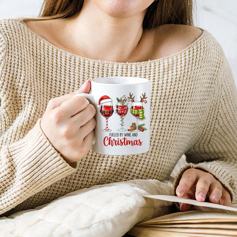 Peut inclure: Mug en c&eacute;ramique blanche avec un motif de No&euml;l. Il pr&eacute;sente trois verres &agrave; vin d&eacute;cor&eacute;s avec des &eacute;l&eacute;ments festifs, dont un bonnet de P&egrave;re No&euml;l et une &eacute;charpe. Le texte "Fueled by Wine and Christmas" est imprim&eacute; en dessous.