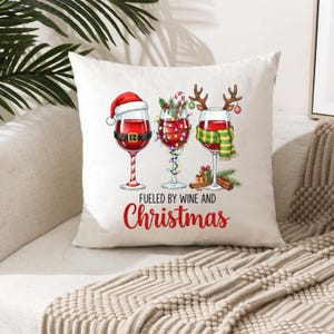 Peut inclure: Un coussin carr&eacute; blanc avec un motif de No&euml;l festif. Il pr&eacute;sente trois verres &agrave; vin d&eacute;cor&eacute;s d'&eacute;l&eacute;ments de vacances, dont un bonnet de P&egrave;re No&euml;l, des guirlandes lumineuses et une &eacute;charpe. Le texte "Fueled by Wine and Christmas" est imprim&eacute; en rouge.