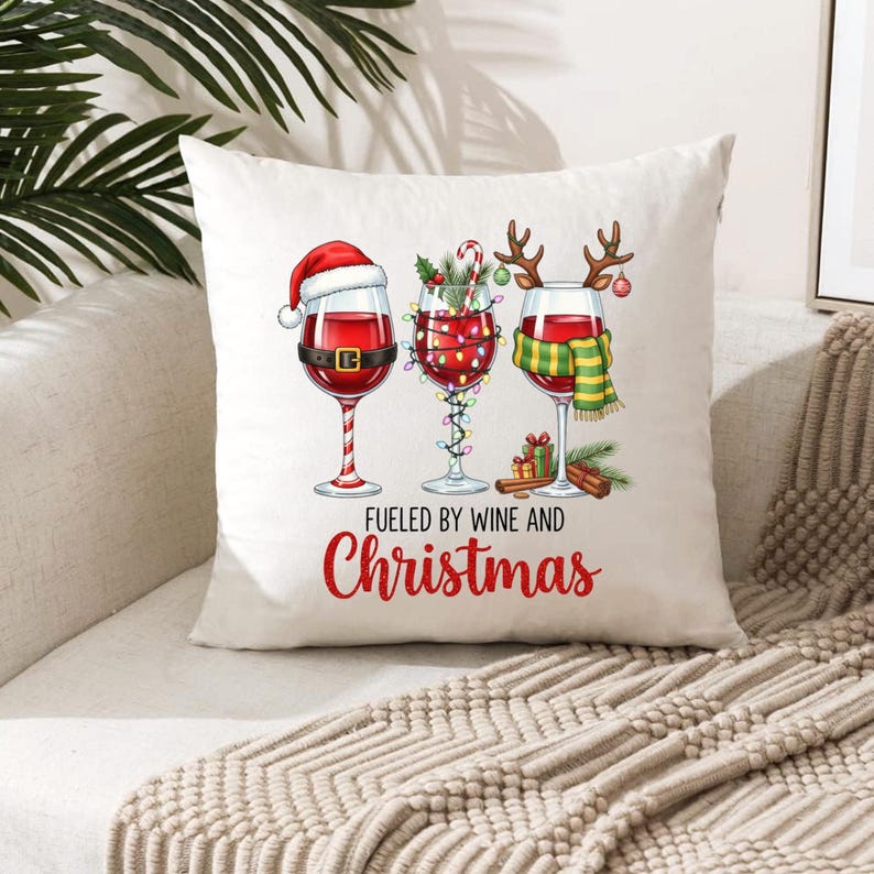 Peut inclure: Un coussin carr&eacute; blanc avec un motif de No&euml;l festif. Il pr&eacute;sente trois verres &agrave; vin d&eacute;cor&eacute;s d'&eacute;l&eacute;ments de vacances, dont un bonnet de P&egrave;re No&euml;l, des guirlandes lumineuses et une &eacute;charpe. Le texte "Fueled by Wine and Christmas" est imprim&eacute; en rouge.