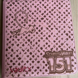 Peut inclure: Classeur rose Vault X avec un motif marron de formes variées et un personnage de dessin animé. Le classeur porte le texte "Scarlet & Violet 151" et le logo Vault X. Conçu pour les cartes à collectionner.
