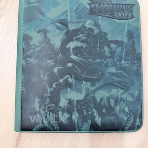 Può includere: Raccoglitore verde Vault X con grafica di drago della serie "Evolving Skies". Il raccoglitore ha una superficie strutturata e il testo "Sword & Shield" e "Evolving Skies" sono visibili. Il raccoglitore è su una superficie di legno chiaro.