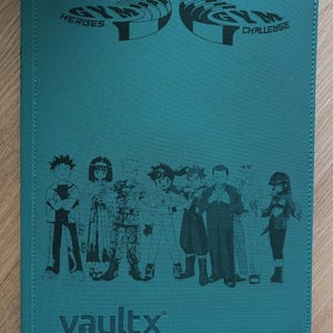 Op de afbeelding: Teal kaartenbinder met de woorden "GYM HEROES" en "GYM CHALLENGE" bovenaan gedrukt. De binder toont een groep cartoonfiguren en het woord "vaultx" onderaan.