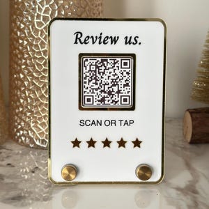 Peut inclure: Un panneau blanc avec une bordure dorée, comportant un code QR et le texte "Review us. SCAN OR TAP". Cinq étoiles dorées se trouvent sous le texte. Deux boutons ronds dorés sont situés en bas du panneau.