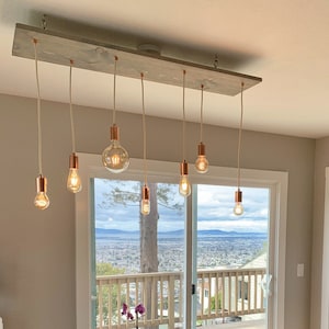 May include: A rustic wood and copper pendant light fixture with six Edison bulbs hanging from a long, weathered wood plank. The fixture is installed in a kitchen with white cabinets and a stainless steel refrigerator.