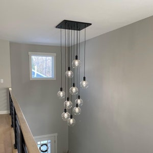 May include: A black metal chandelier with ten clear glass globe light bulbs hanging from a square metal base. The chandelier is hanging from the ceiling in a room with a wooden staircase.