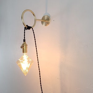 May include: A gold wall sconce with a circular ring and a black cord hanging down. A diamond-shaped light bulb is attached to the cord.