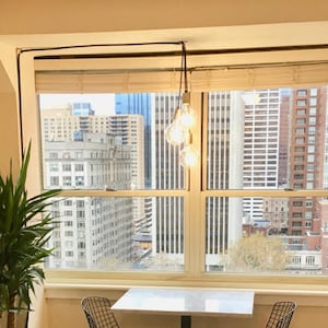 Peut inclure: Une table blanche avec un cadre en métal noir et deux chaises en fil de fer noir sont placées devant une grande fenêtre offrant une vue sur un horizon urbain. Un luminaire suspendu avec quatre ampoules apparentes est suspendu au-dessus de la table.