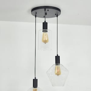 Puede incluir: Una lámpara de techo negra con tres luces colgantes. Cada luz tiene una pantalla de vidrio transparente de forma diferente y una bombilla de luz blanca cálida.