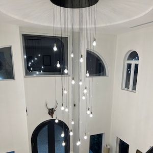 Może przedstawiać: Duży, nowoczesny żyrandol z wieloma żarówkami wiszącymi na czarnej metalowej ramie. Żyrandol jest zawieszony na suficie domu dwupiętrowego z klatką schodową.