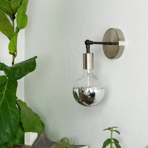 Puede incluir: Una lámpara de pared plateada con una bombilla de globo de vidrio transparente. La lámpara está montada en una pared blanca junto a una planta verde. Una pequeña mesa de madera con libros y una planta en un frasco de vidrio se encuentra frente a una almohada verde.