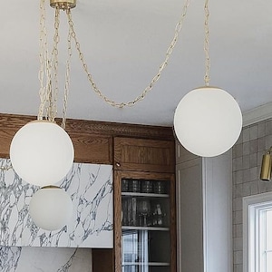 Peut inclure: Un îlot de cuisine avec un comptoir noir et quatre tabourets de bar noirs et bruns. L'îlot est devant une crédence en marbre blanc et des armoires en bois. Trois lampes suspendues blanches en forme de globe sont suspendues au-dessus de l'îlot.