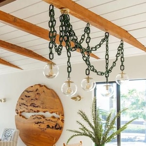 May include: A ceiling-mounted chandelier featuring five clear glass globe light bulbs suspended by dark green plastic chains that drape across wooden ceiling beams. The fixture includes brass hardware connecting the chains to the glass globes.