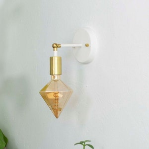 Könnte beinhalten: Eine weiße Wandleuchte mit einem goldenen Arm und einer diamantförmigen Glühbirne. Das Licht ist an und beleuchtet den Raum. Auf einem Tisch unter der Leuchte stehen Bücher und Pflanzen.