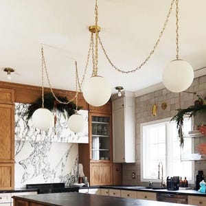 May include: A kitchen interior featuring three pendant lights with spherical, white glass shades suspended from gold chains. The kitchen has wooden cabinets, a marble backsplash, and a dark countertop island. Decorative greenery adds a natural touch to the space.