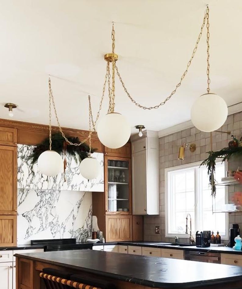 May include: A kitchen interior featuring three pendant lights with spherical, white glass shades suspended from gold chains. The kitchen has wooden cabinets, a marble backsplash, and a dark countertop island. Decorative greenery adds a natural touch to the space.