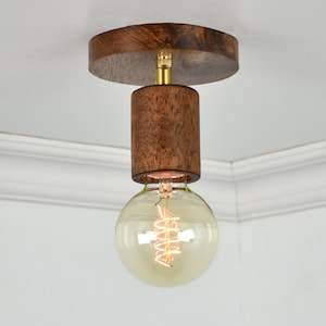 May include: A wooden ceiling light fixture with a round canopy and a cylindrical shade. The light fixture has a brass finish and a clear glass bulb with a coiled filament.