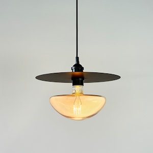 Könnte beinhalten: Eine schwarze Pendelleuchte mit einem runden, bernsteinfarbenen Glasschirm und einer schwarzen Metallscheibe über der Glühbirne.