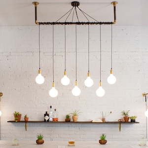 K&ouml;nnte beinhalten: Eine moderne Leuchte im Industriestil mit acht Edison-Gl&uuml;hbirnen, die an einem schwarzen Rohr und Kabel h&auml;ngen. Die Leuchte ist an einer wei&szlig;en Backsteinwand montiert.