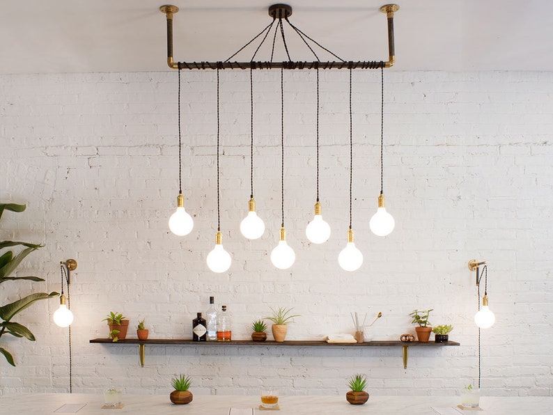 K&ouml;nnte beinhalten: Eine moderne Leuchte im Industriestil mit acht Edison-Gl&uuml;hbirnen, die an einem schwarzen Rohr und Kabel h&auml;ngen. Die Leuchte ist an einer wei&szlig;en Backsteinwand montiert.