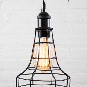 May include: Black pendant light with a cage-style shade and a vintage-style light bulb. The light is hanging from a black cord against a white brick wall.