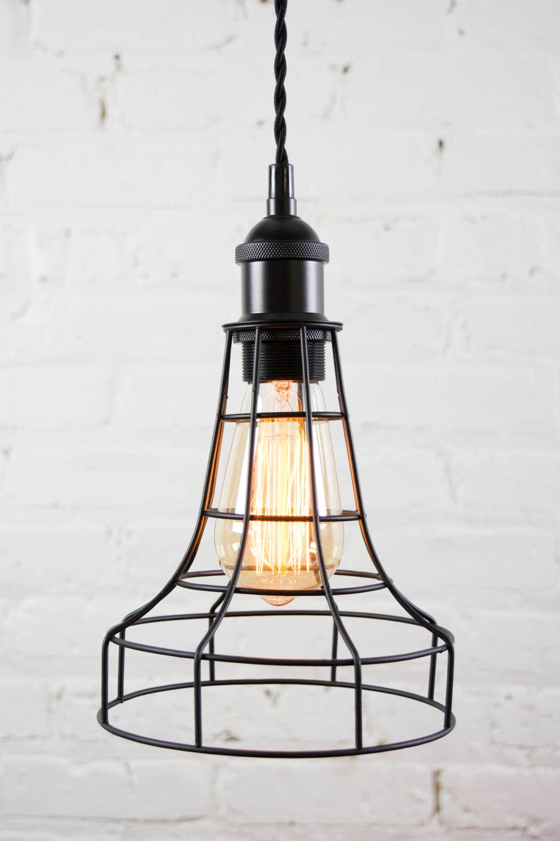 May include: Black pendant light with a cage-style shade and a vintage-style light bulb. The light is hanging from a black cord against a white brick wall.