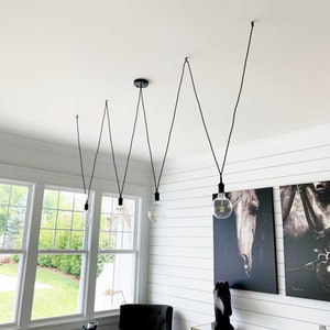 May include: Five black pendant lights with clear glass bulbs hang from a white ceiling in a dining room. The lights are suspended from black cords and are arranged in a staggered pattern. The room has white shiplap walls and a large window with a view of a green lawn.