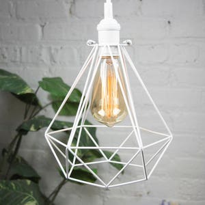 Puede incluir: L&aacute;mpara colgante geom&eacute;trica blanca con una bombilla de vidrio transparente. La luz cuelga de un cable blanco contra una pared de ladrillo blanco.