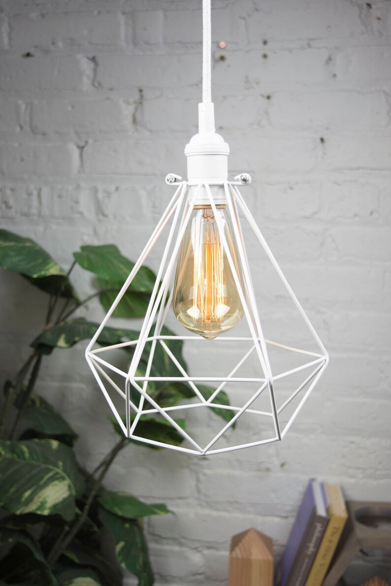 Puede incluir: L&aacute;mpara colgante geom&eacute;trica blanca con una bombilla de vidrio transparente. La luz cuelga de un cable blanco contra una pared de ladrillo blanco.