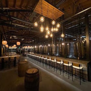 Puede incluir: Una barra de madera larga con taburetes de bar blancos y una fila de barriles marrones en primer plano. La barra est&aacute; iluminada por una serie de bombillas colgantes.