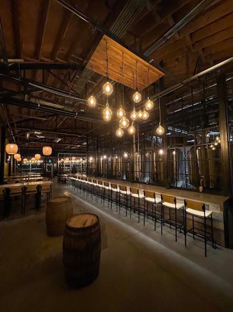 Puede incluir: Una barra de madera larga con taburetes de bar blancos y una fila de barriles marrones en primer plano. La barra est&aacute; iluminada por una serie de bombillas colgantes.