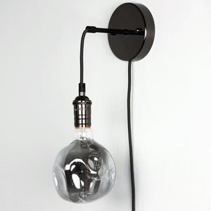 K&ouml;nnte beinhalten: Eine schwarze Wandleuchte mit runder Basis und schwarzem Kabel. Eine klare Gl&uuml;hbirne mit strukturierter Oberfl&auml;che ist am Kabel befestigt.