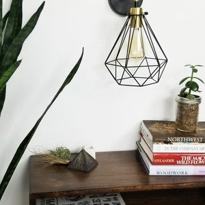 May include: A modern wall sconce with a black geometric shade and a gold-toned bulb holder. The sconce is mounted on a dark circular base. A wooden shelf below holds books, a small plant, and decorative items. The overall theme is modern home decor.