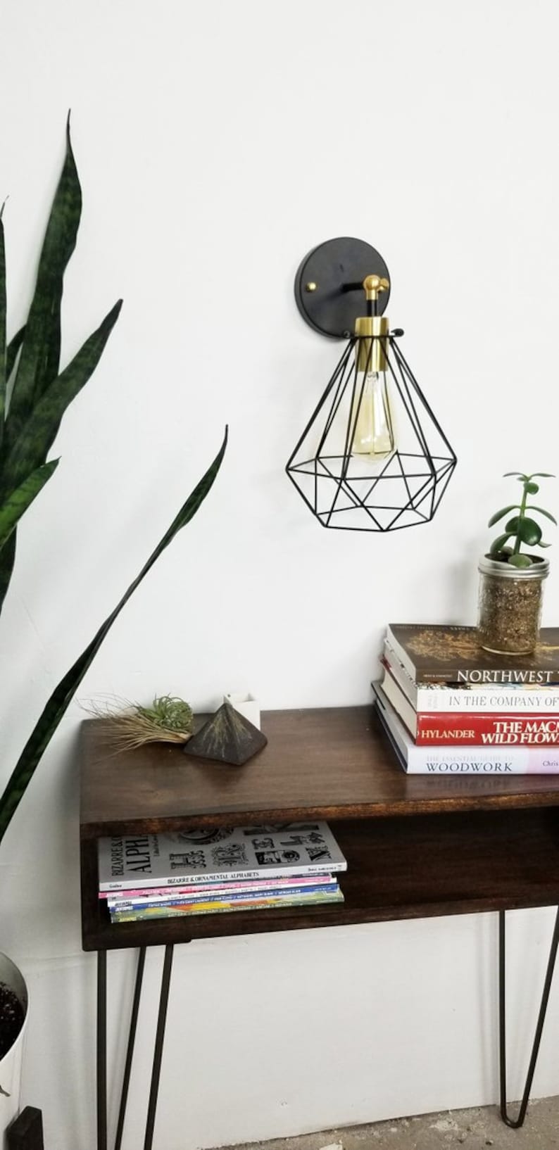 May include: A modern wall sconce with a black geometric shade and a gold-toned bulb holder. The sconce is mounted on a dark circular base. A wooden shelf below holds books, a small plant, and decorative items. The overall theme is modern home decor.