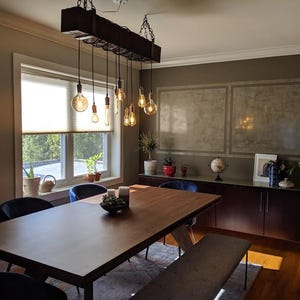 May include: A wooden dining table with a rustic, industrial style chandelier hanging above it. The table is surrounded by blue chairs and a bench. The room has a modern, minimalist design with a concrete wall and a large window.