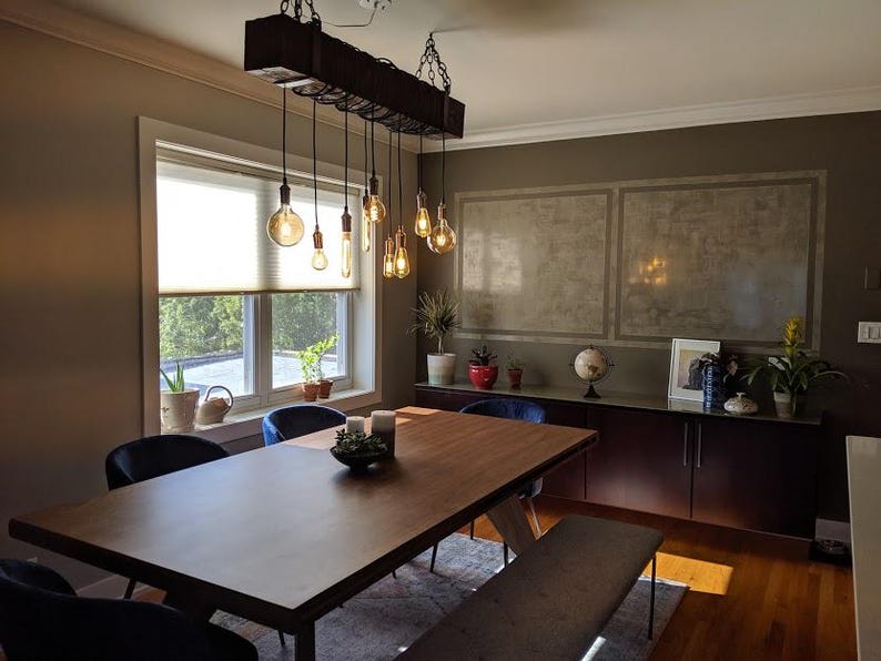 May include: A wooden dining table with a rustic, industrial style chandelier hanging above it. The table is surrounded by blue chairs and a bench. The room has a modern, minimalist design with a concrete wall and a large window.