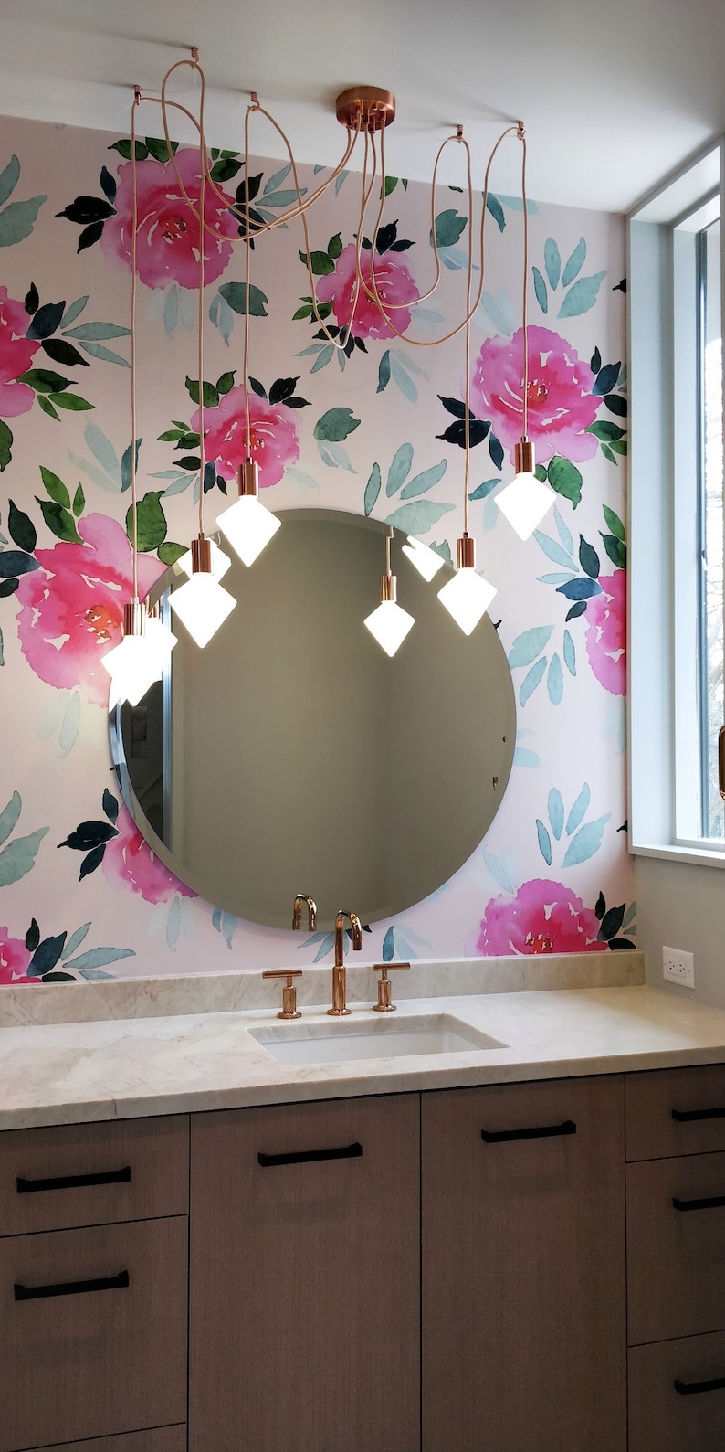 K&ouml;nnte beinhalten: Ein Badezimmer-Waschtisch mit einem runden Spiegel und einer floralen Tapete mit rosa Blumen und gr&uuml;nen Bl&auml;ttern. Der Waschtisch hat eine wei&szlig;e Arbeitsplatte mit einem Waschbecken und einem Satz Kupferarmaturen. Der Spiegel wird von einem Kronleuchter mit sechs Gl&uuml;hbirnen beleuchtet.