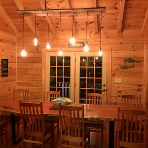 May include: A rustic dining room with a wooden table and chairs. The table is made of a live edge slab of wood and has a natural finish. The chairs are made of wood and have a simple design. The room has a wooden ceiling and walls, and there is a chandelier with Edison bulbs hanging from the ceiling. There is a sign on the wall that says "Life is better at the lake".