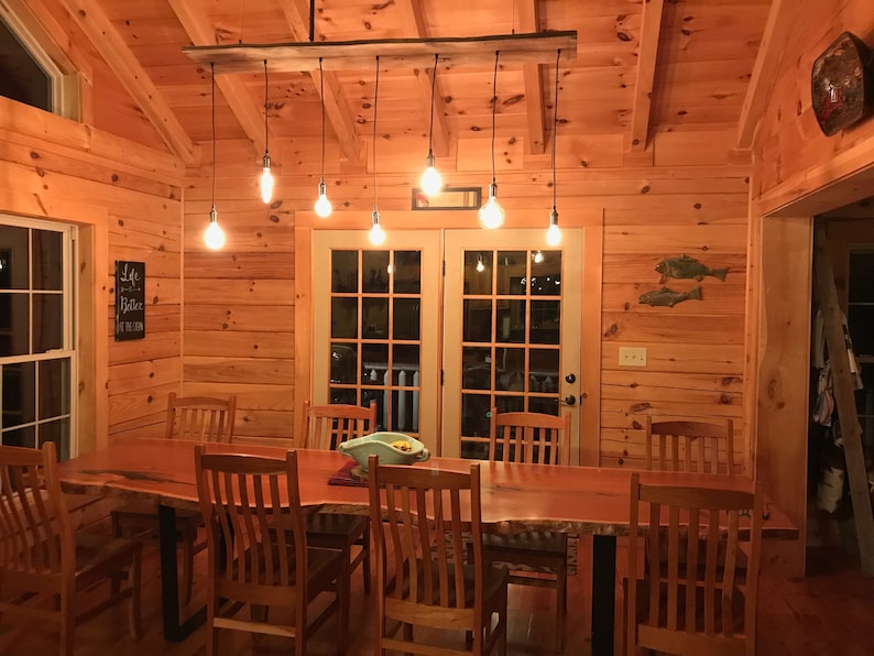 May include: A rustic dining room with a wooden table and chairs. The table is made of a live edge slab of wood and has a natural finish. The chairs are made of wood and have a simple design. The room has a wooden ceiling and walls, and there is a chandelier with Edison bulbs hanging from the ceiling. There is a sign on the wall that says "Life is better at the lake".