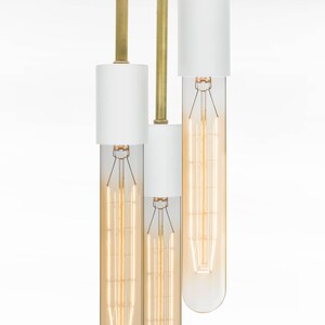 May include: A white ceiling light fixture with three brass rods and white cylindrical shades. Each shade has a clear glass bulb with a filament inside.