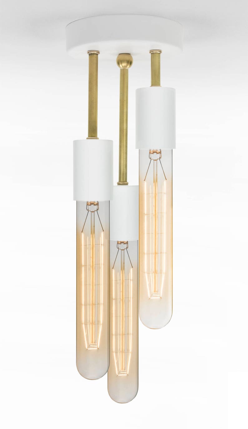May include: A white ceiling light fixture with three brass rods and white cylindrical shades. Each shade has a clear glass bulb with a filament inside.