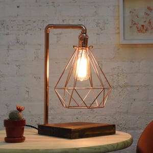 Puede incluir: Una l&aacute;mpara de mesa de cobre con una pantalla de alambre geom&eacute;trico y una base de madera. La l&aacute;mpara est&aacute; encendida y la bombilla ilumina la pantalla.