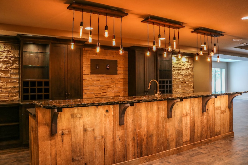 May include: A rustic wooden bar with a granite countertop and a dark brown wood cabinet behind it. The bar is lit by multiple hanging Edison bulb lights.