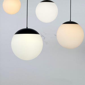 May include: Four modern pendant lights with spherical white glass shades and black caps. The lights hang from black cords against a light gray background. The shades emit a soft, warm glow, creating a minimalist aesthetic.