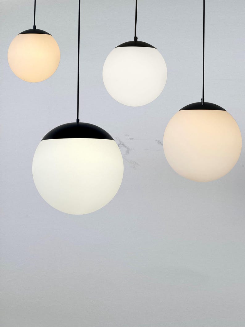 May include: Four modern pendant lights with spherical white glass shades and black caps. The lights hang from black cords against a light gray background. The shades emit a soft, warm glow, creating a minimalist aesthetic.