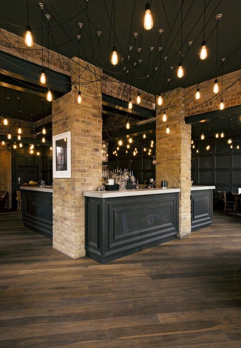 May include: A dark wood bar with a black countertop and black cabinets. The bar is lit by a series of exposed Edison bulbs hanging from the ceiling. The walls are made of exposed brick.