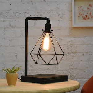 Puede incluir: Una lámpara de mesa de metal negro con una pantalla geométrica en forma de diamante. La lámpara tiene una base de metal negro y un cable negro.
