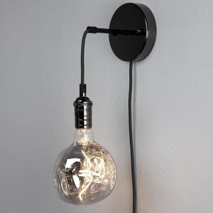 K&ouml;nnte beinhalten: Eine schwarze Wandleuchte mit runder Basis und einem langen Kabel. Eine klare Gl&uuml;hbirne mit Filamentdesign ist am Kabel befestigt.