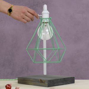 Puede incluir: Una pantalla de l&aacute;mpara de jaula de alambre verde con una base blanca y una base de madera. La l&aacute;mpara est&aacute; encendida y la bombilla es visible.