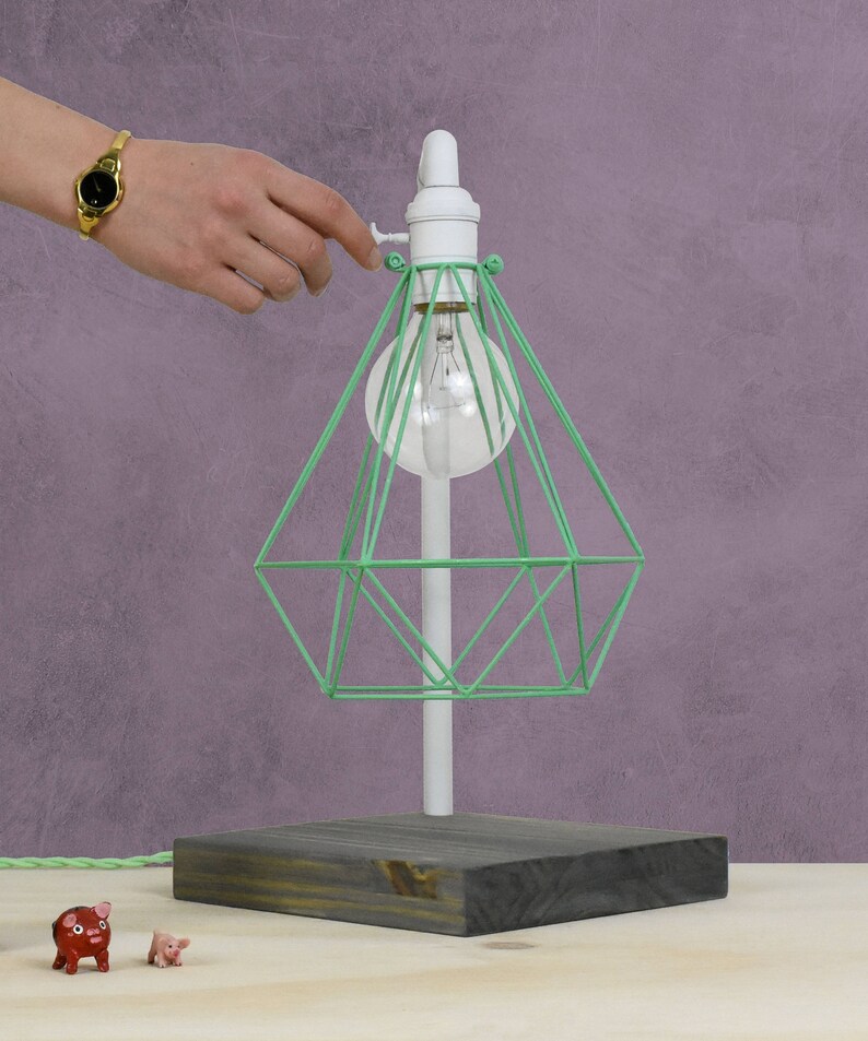 Puede incluir: Una pantalla de l&aacute;mpara de jaula de alambre verde con una base blanca y una base de madera. La l&aacute;mpara est&aacute; encendida y la bombilla es visible.